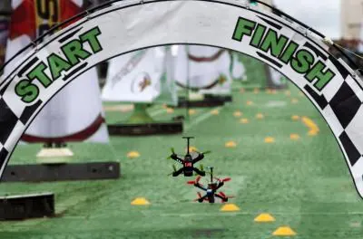 Two small racing drones flying through a start and finish gate over artificial turf