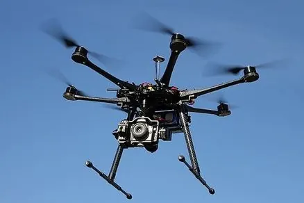 Black hexacopter drone with six rotors carrying a camera under its frame against a blue sky