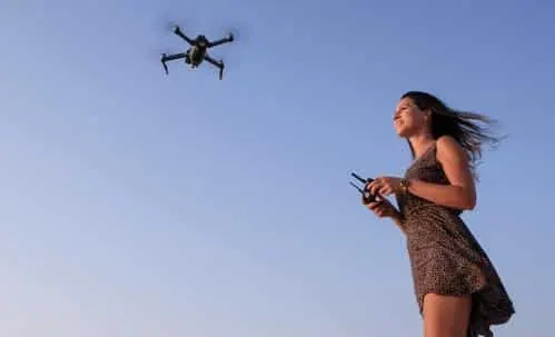 Woman holding a transmitter and flying a black drone overhead against a blue sky