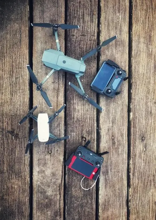 Two folding drones, a white mini drone and their controllers laid out on a wooden deck