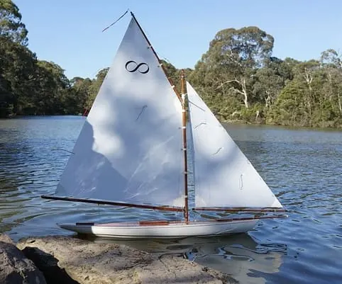 White RC sailboat with two sails on a lake surrounded by trees