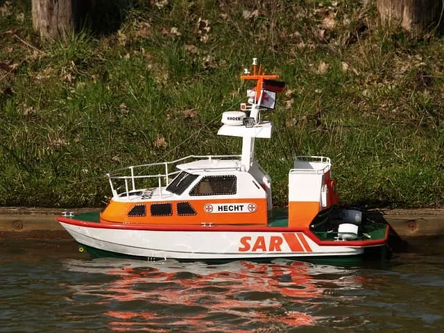 Orange and white RC rescue boat with a cabin and a mast, floating on calm water next to a bank